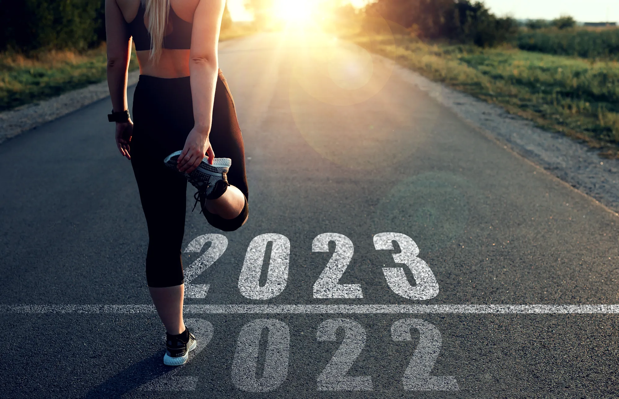 A female runner stands at the finish line of 2022 and the starting line of 2023 stretching, while facing down the road toward the rising sun of the new year. Presented by Miami Beach Brokers®