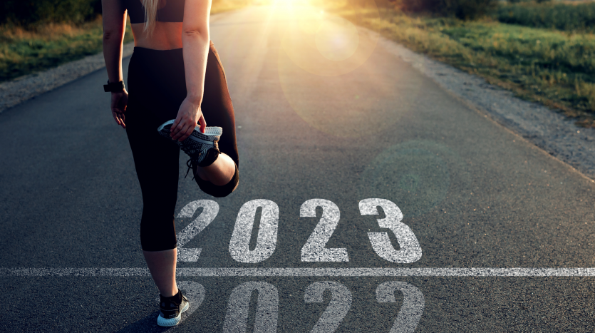 A female runner stands at the finish line of 2022 and the starting line of 2023 stretching, while facing down the road toward the rising sun of the new year. Presented by Miami Beach Brokers®