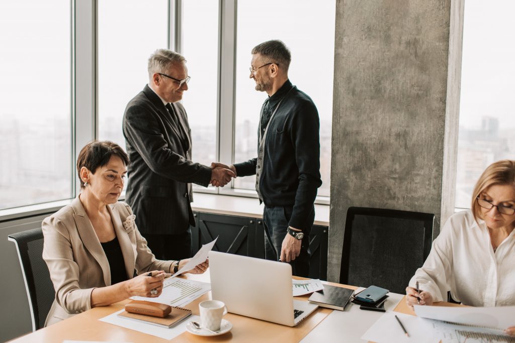 Four professionals in modern office; two shaking hands near large windows.